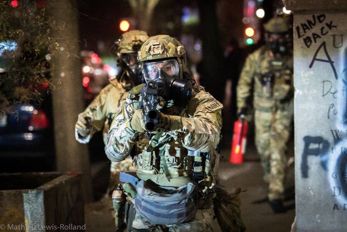 Two armed soldiers with gas masks on, a third is seen in the background with a fire extinguisher. One points his weapon at the camera.