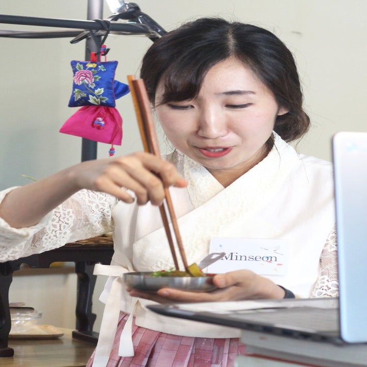 Minseon, the host, using chopsticks to stir something in a small bowl in front of a laptop screem