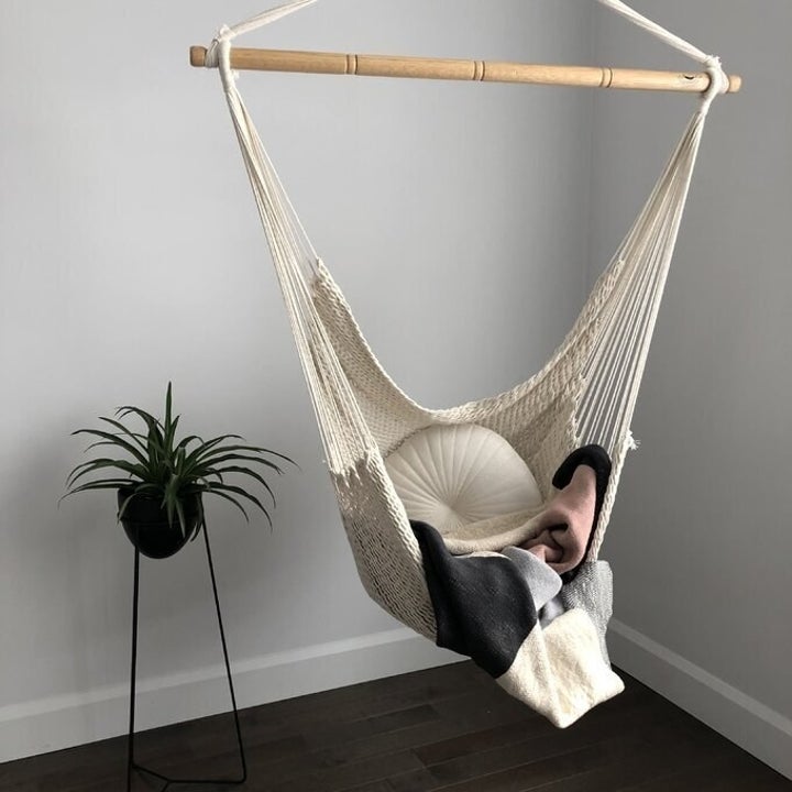 A reviewer image of their hammock in a corner of their home, filled with a pillow and decorative blanket and placed beside a potted plant