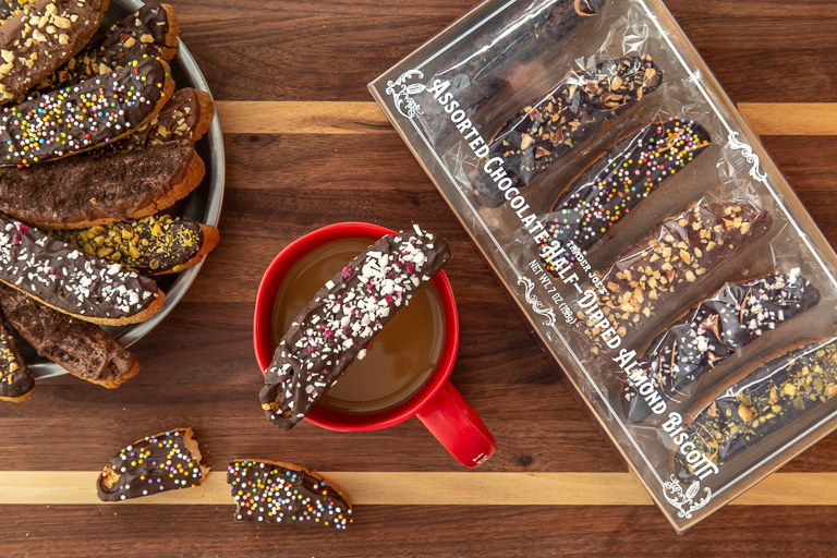 A cup of coffee sits upon a wooden table. On top of the cup balances a chocolate biscotti with candy cane pieces baked in. The coffee cup is surrounded by biscotti cookies.