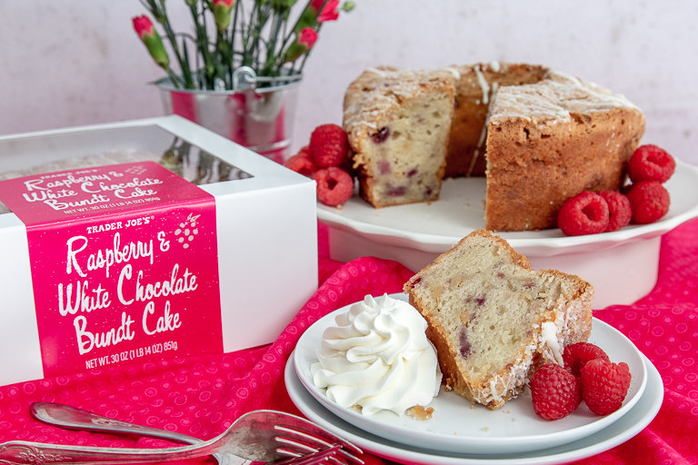 A box that reads, &quot;Raspberry white chocolate bundt cake&quot; sits on a tablecloth next to two plates. One large plate contains the bundt cake surrounded by raspberries, and one slice is taken out. The cut slice sits on another plate, next to whipped cream