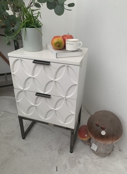 A carved nightstand with circles and diamond shapes on each drawer. There are two drawers that can be pulled open with flat metal handles that match the square legs of the dresser 