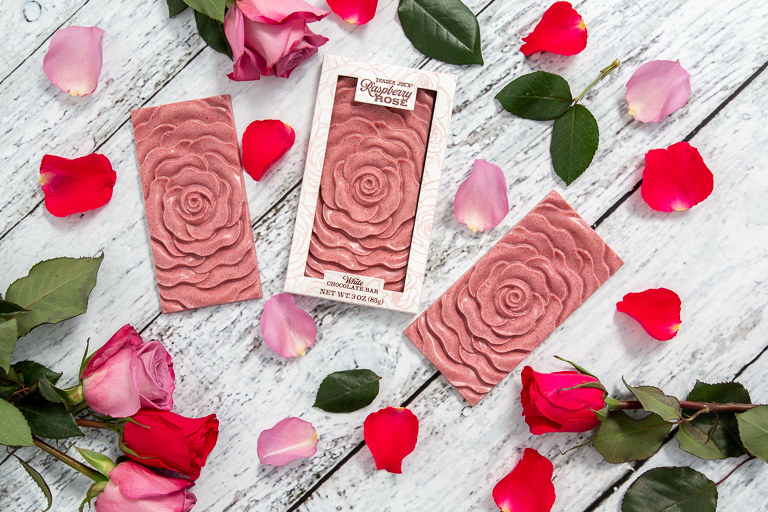 Rectangular chocolate bars with rose designs sit upon a wooden table. Surrounding the chocolate bars are rose petals.