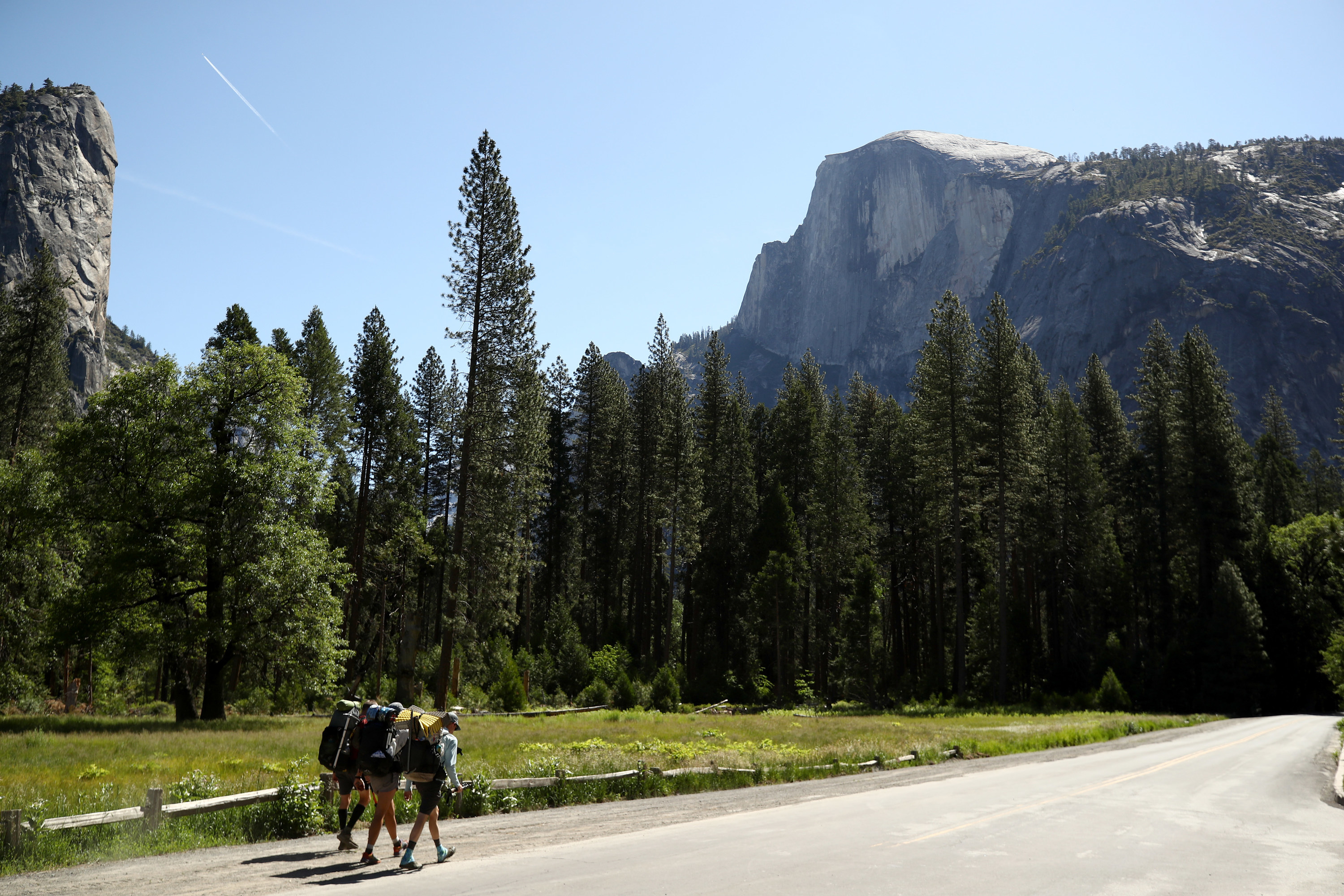 Three people wearing rucksacks hike along an empty road amid tall trees and a steep mountain in the background