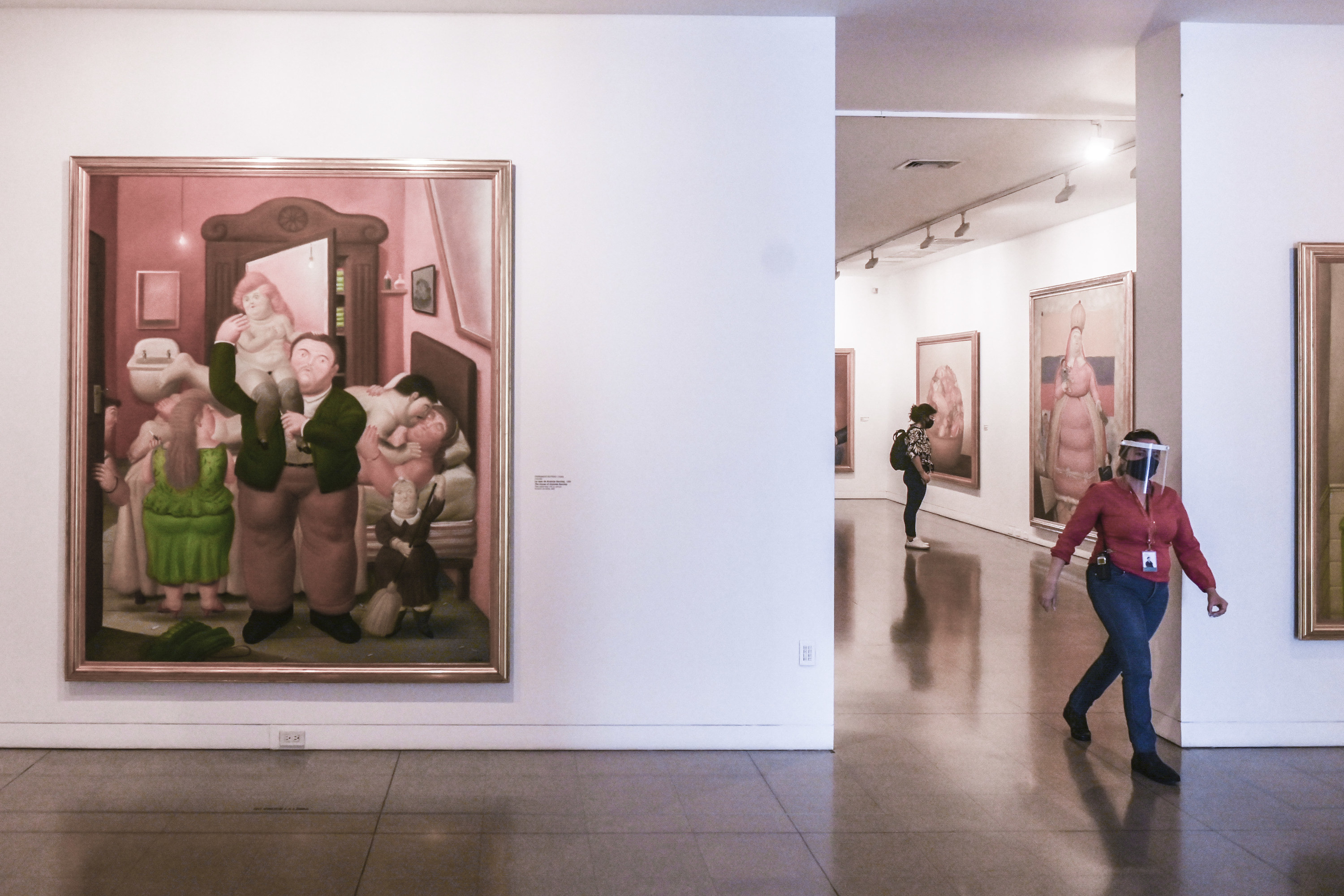 A person wearing a face mask and shield walks between rooms in an art gallery where large paintings are framed on the walls and only one other visitor is seen in the background