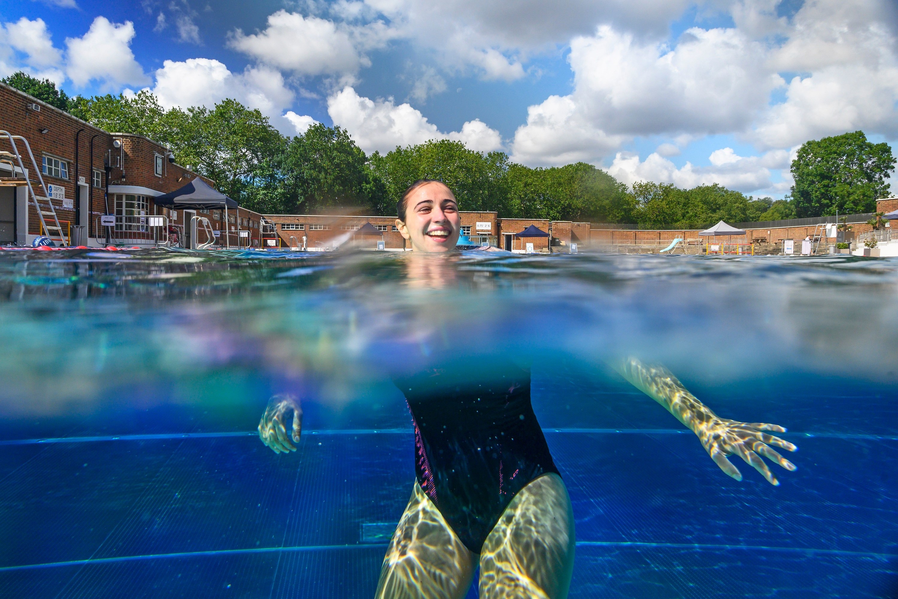 A woman wearing a bathing suit swims in a pool and smiles with the camera angle half above the water and half below