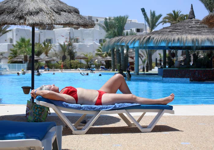 A woman in a bikini lounges on a deck chair under a thatch umbrella next to a hotel pool where people are swimming