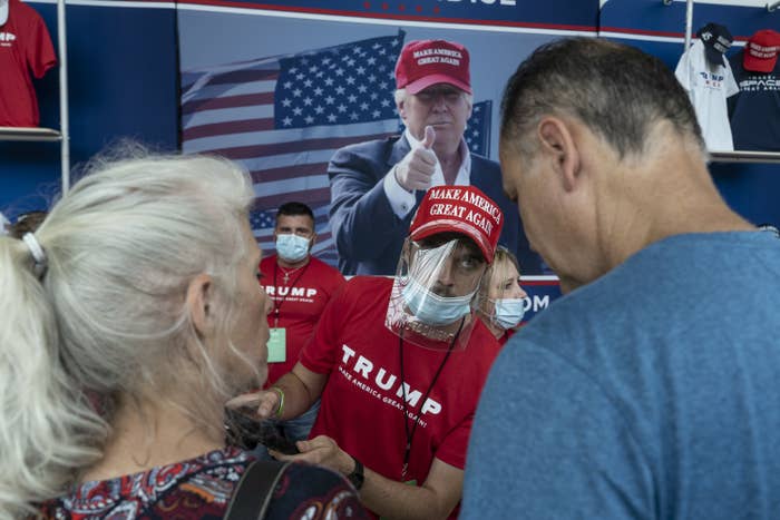 A vendor at a rally wears a &quot;Make America Great Again&quot; hat and T-shirt, protective face shield, and face mask