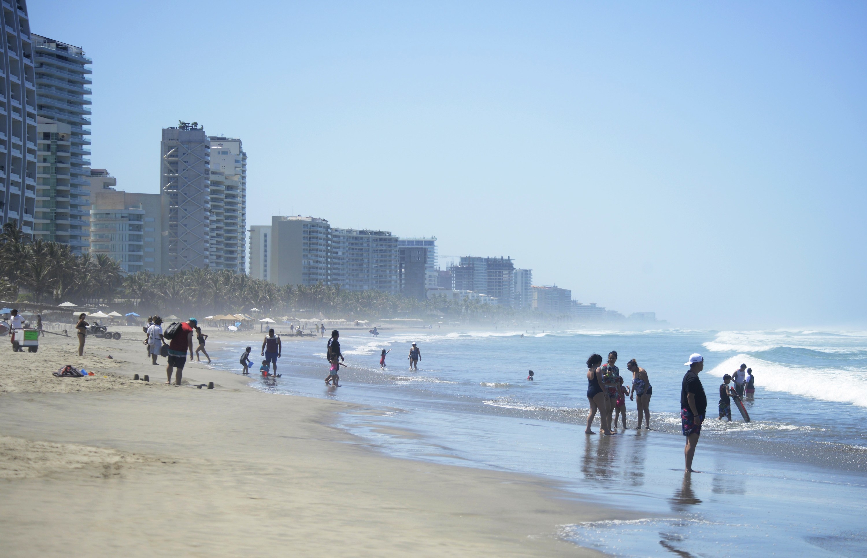 People stand on a beach lined with high-rise apartment blocks as waves crash on the shore under a sunny sky