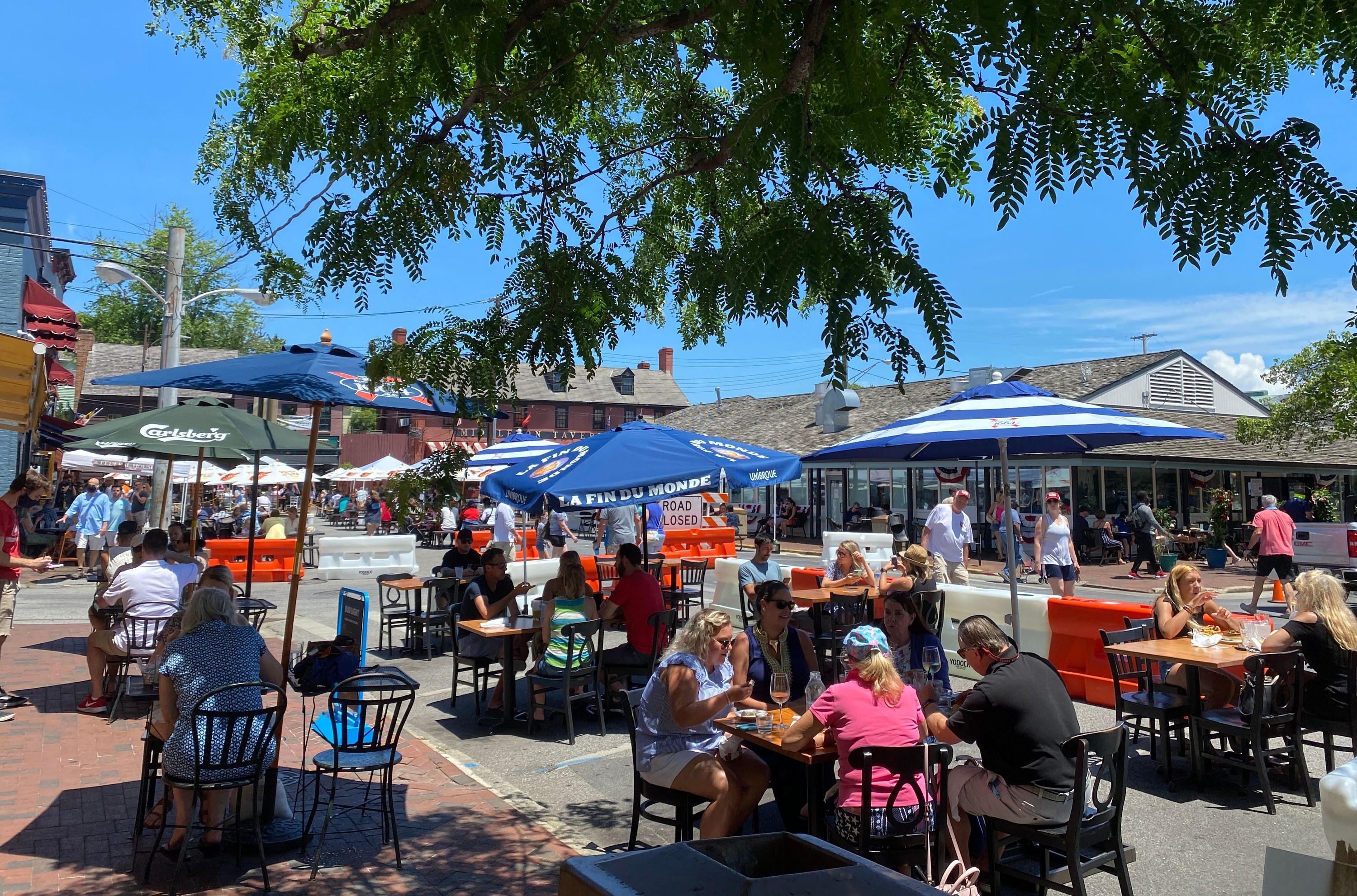 People sit outside at chairs and tables spaced apart under umbrellas as they dine under sunny skies