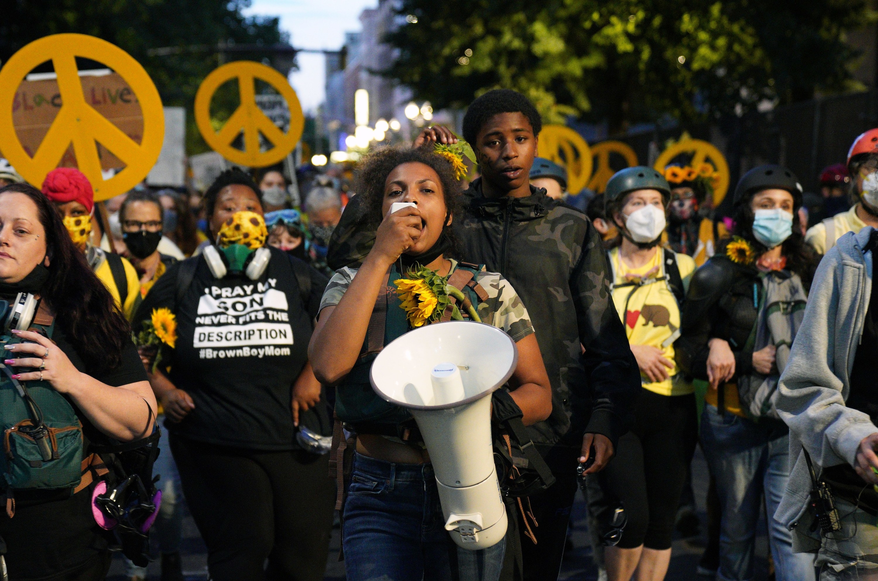 A young Black woman protester speaks into a megaphone in front of a group of demonstrators carrying peace signs
