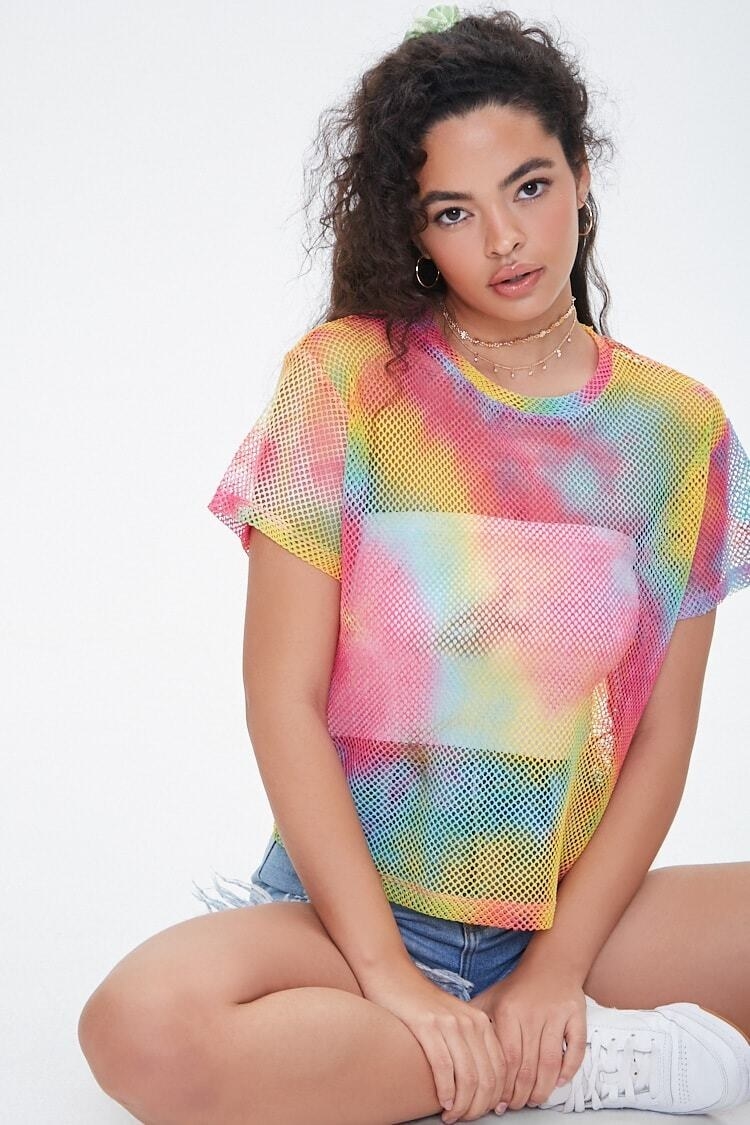 A model wearing the mesh shirt over a tight crop top. The shirt has a boxy fit with a standard crewneck collar and a watercolor-like rainbow and white tie dye pattern throughout. 