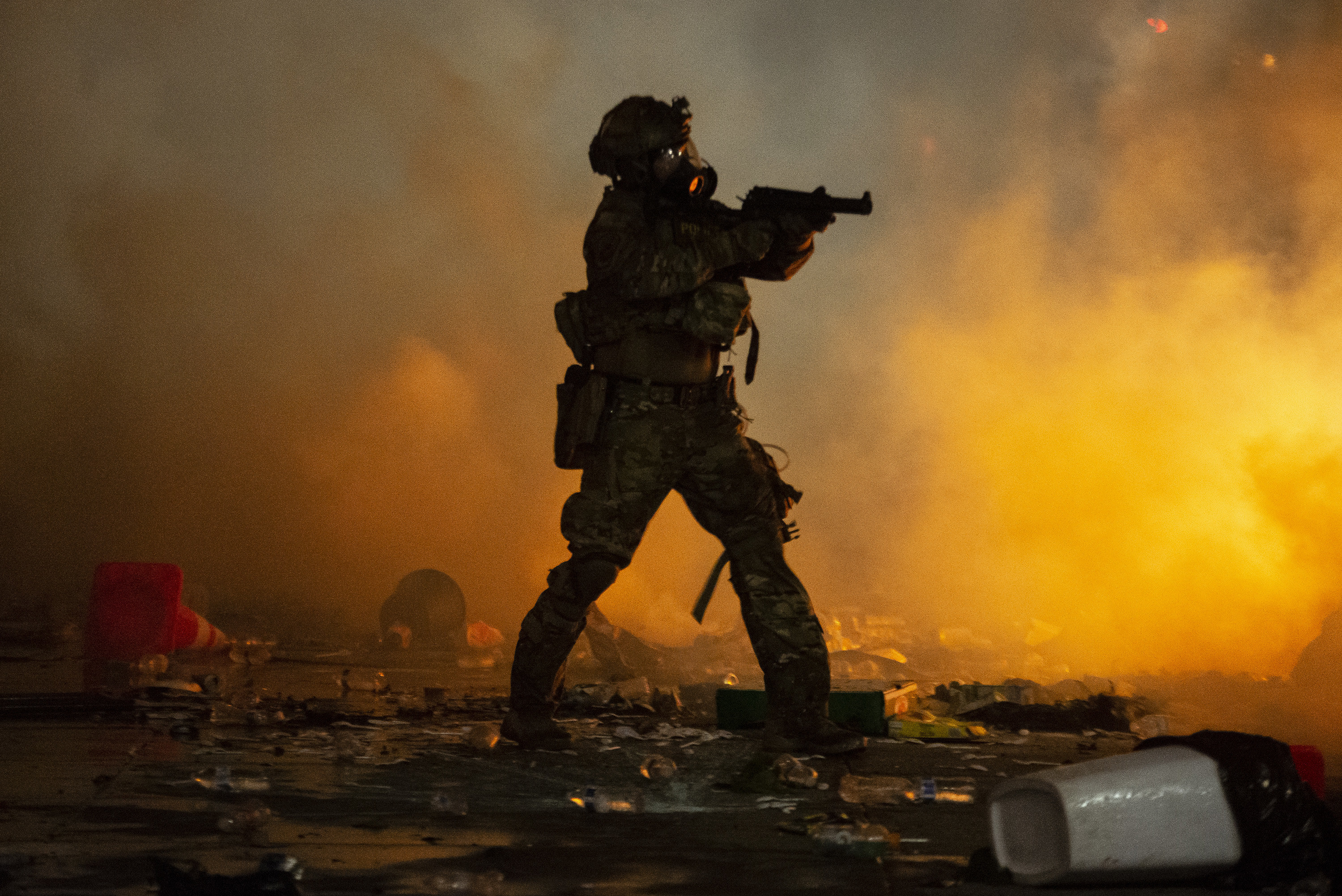 A federal officer points a weapon that looks like a gun but is used to fire less-lethal crowd control mechanisms towards a crowd that is unseen due to tear gas