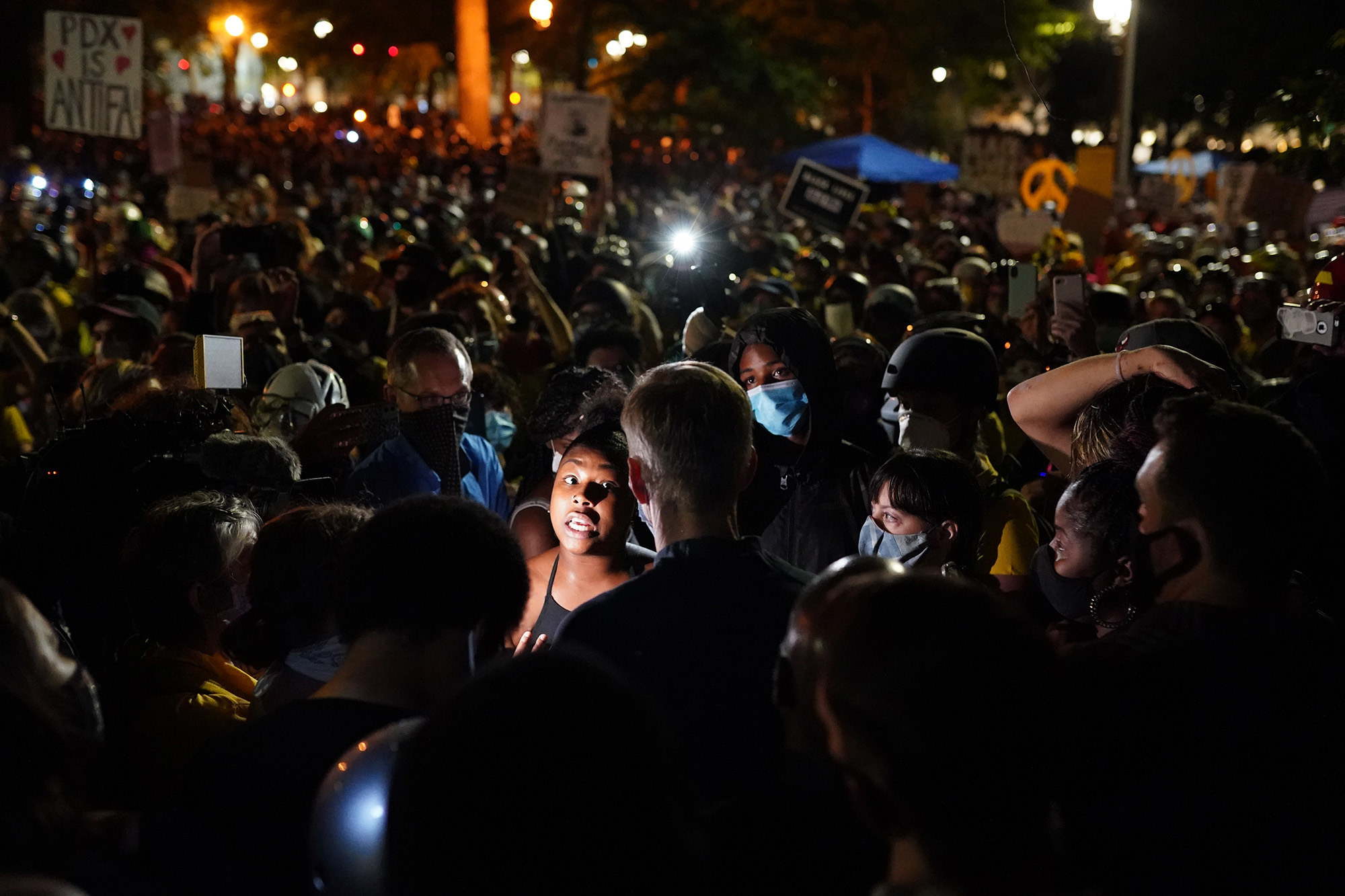 A protester illuminated by a camera flash speaks with Portland Mayor Ted Wheeler surrounded by a large crowd