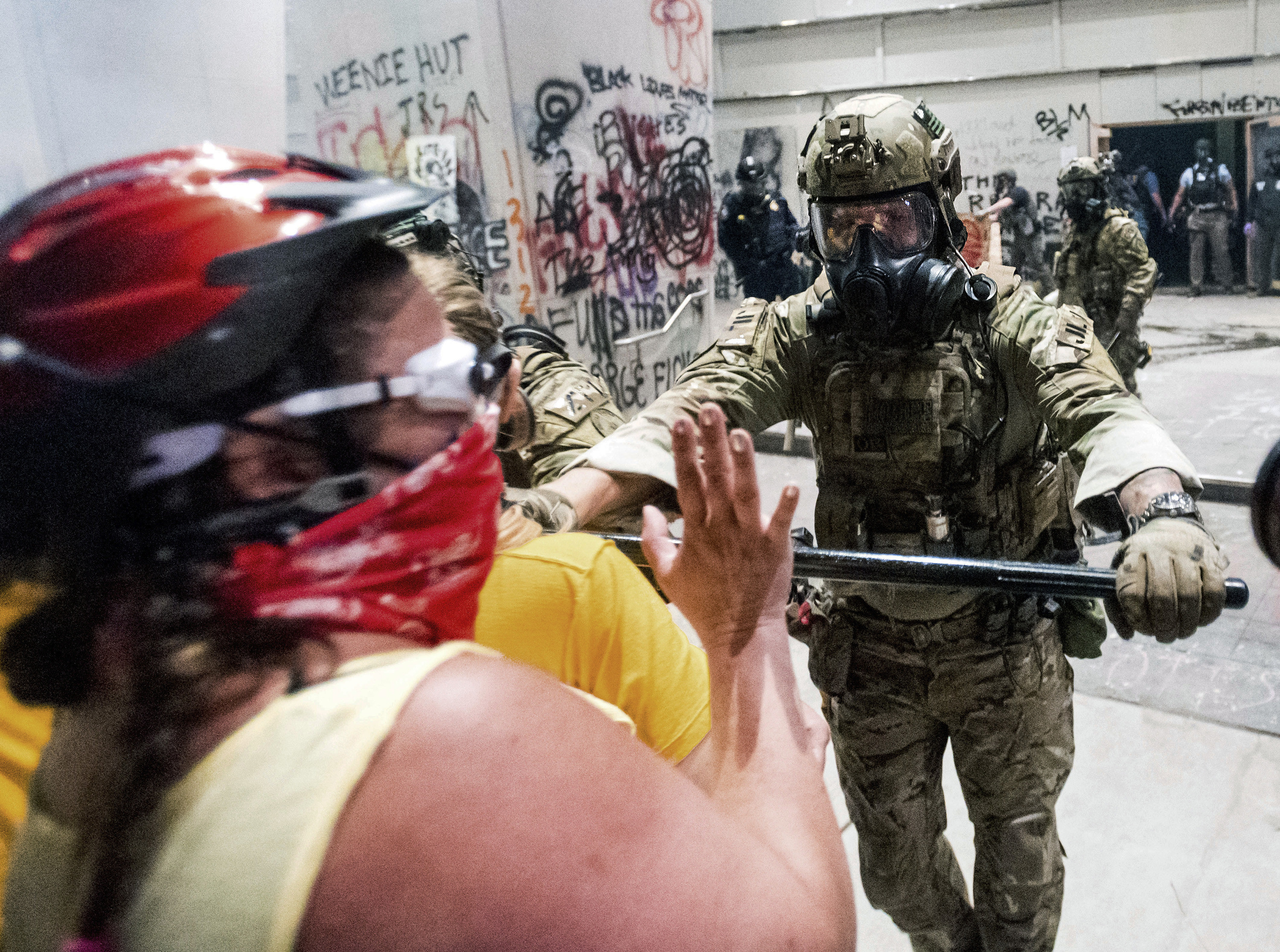 A federal officer in camouflage, a helmet, and gas mask uses a baton to push back women wearing matching yellow shirts