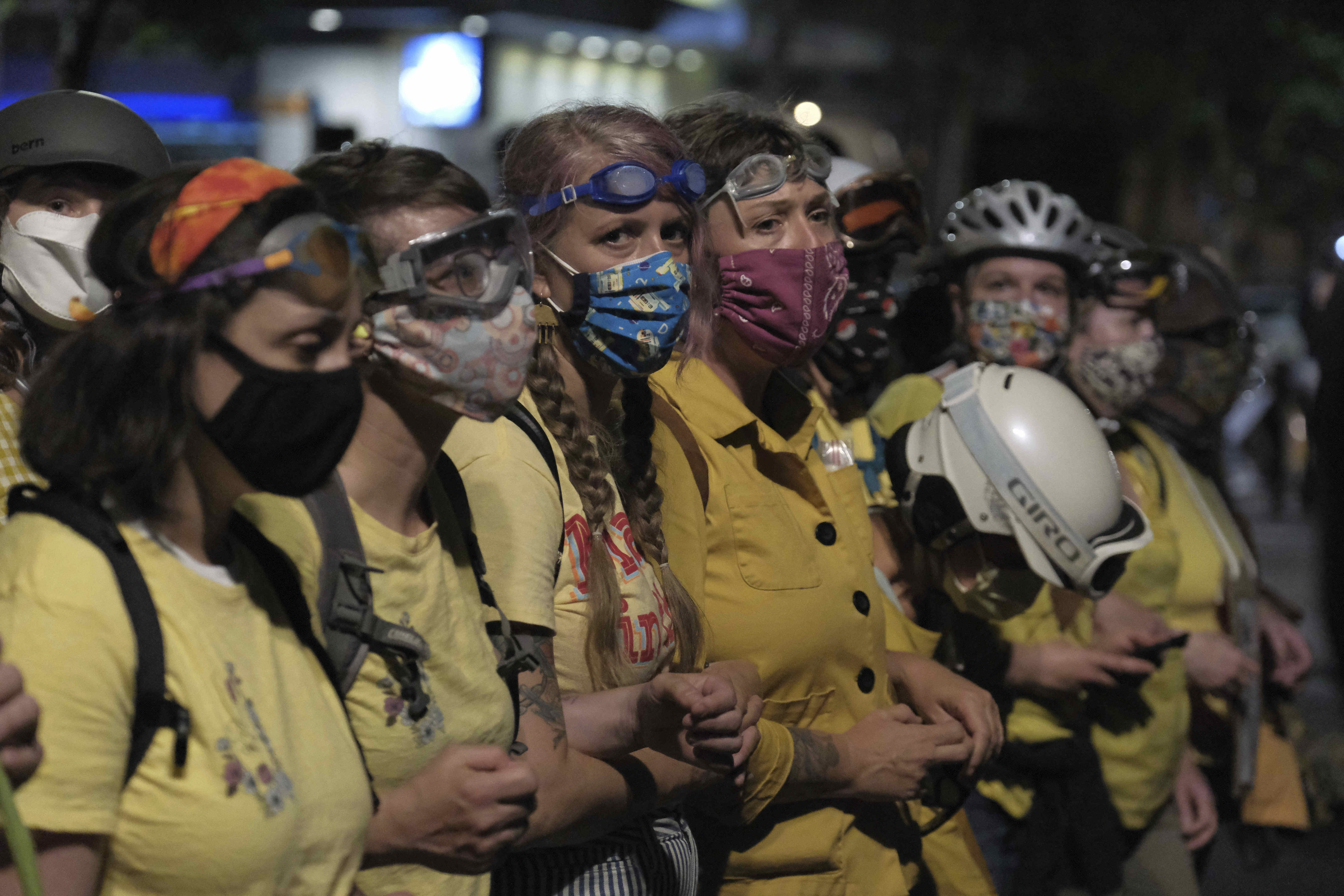 Moms in yellow shirts with interlocking arms stand between protesters and a fence blocking access to the entrance of the federal courthouse