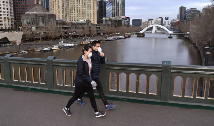 A man and woman wearing face masks and puffy jackets walk across a bridge on a river with tall city buildings in the background.