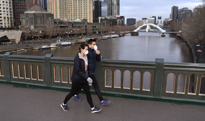 A man and woman wearing face masks and puffy jackets walk across a bridge on a river with tall city buildings in the background. 