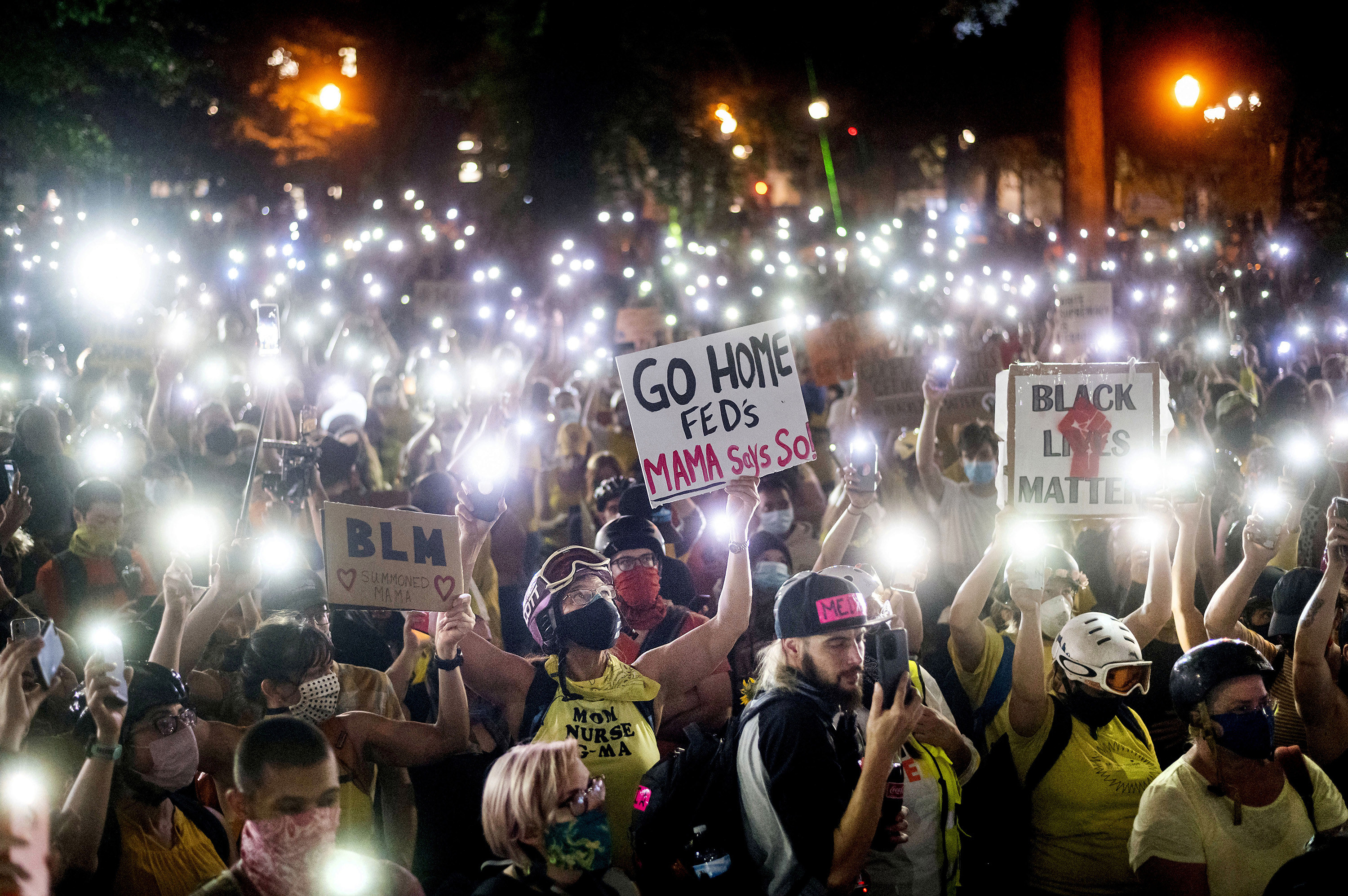 Hundreds of Black Lives Matter protesters hold their phones aloft creating a sea of flashlights. Signs in the crowd read &quot;BLM&quot; and &quot;Go Home Feds. Mama says so!&quot;