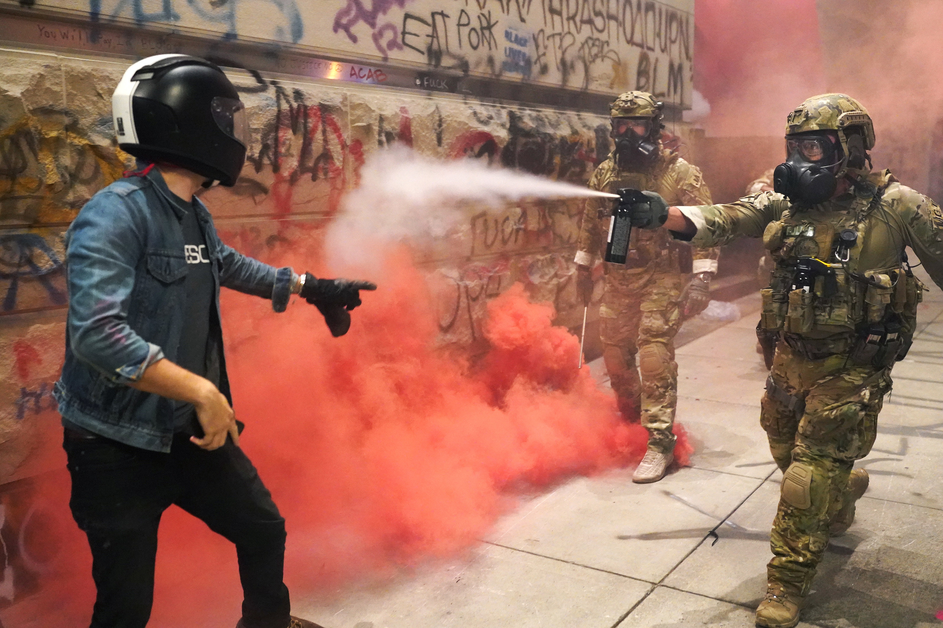 Two federal officers in camouflage use pepper spray on a protester wearing a bike helmet