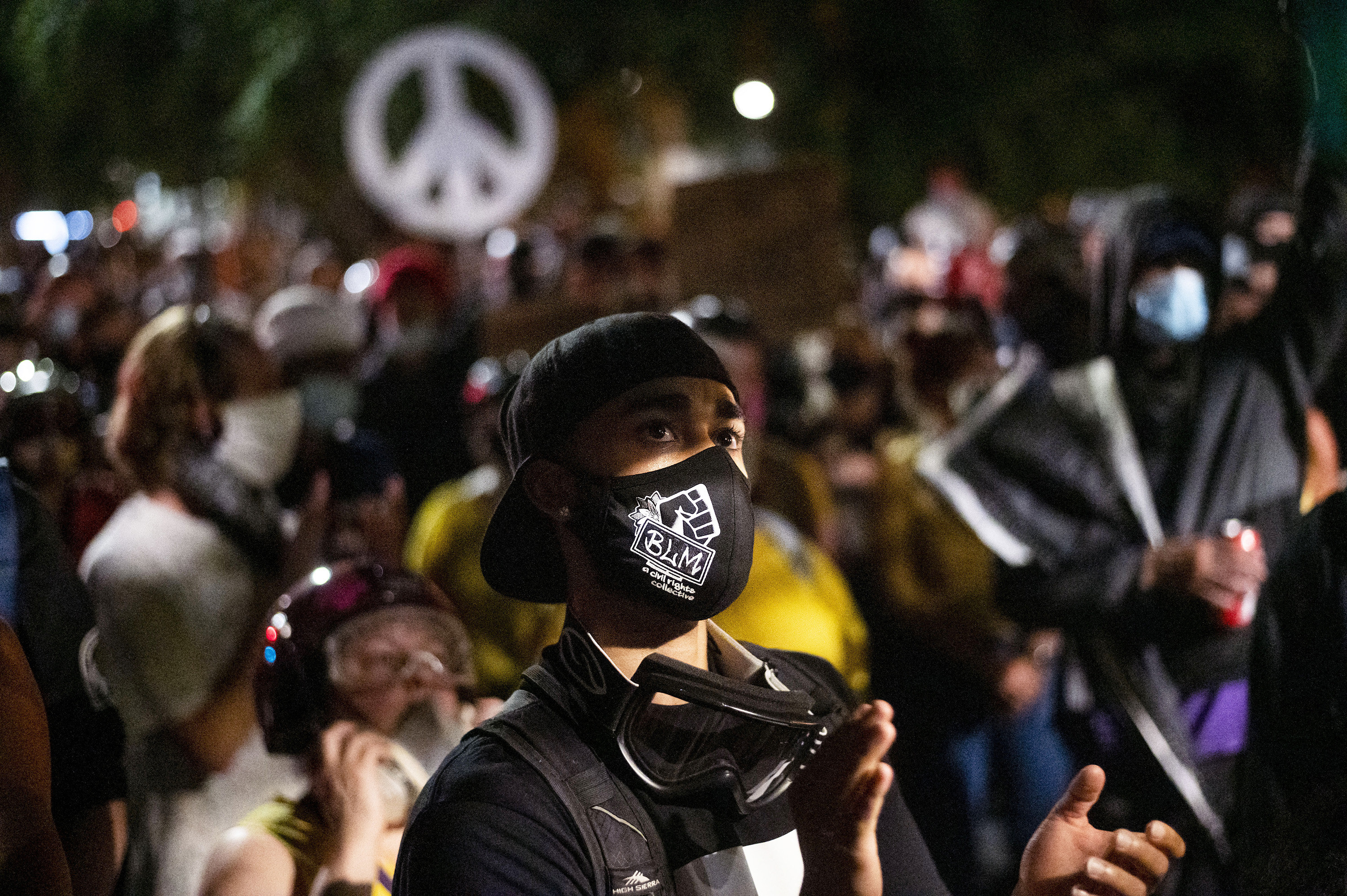 A protester in a black mask that reads &quot;BLM&quot; applauds as they stare off camera