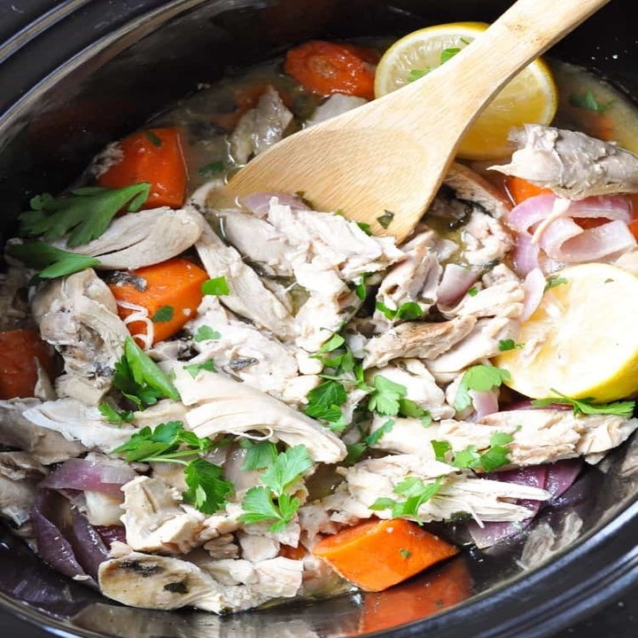 A wooden spoon stirring shredded chicken with carrots, lemon, and herbs in a slow cooker.