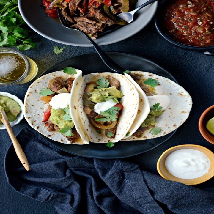 Four steak fajitas topped with guacamole and sour cream, surrounded by more fajita ingredients.