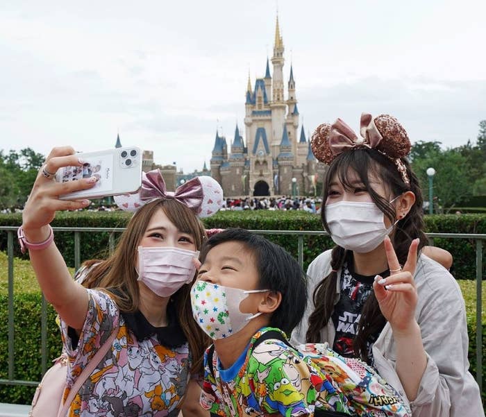 Three people pose for a selfie wearing face masks and ears in front of the Disneyland castle in Tokyo.
