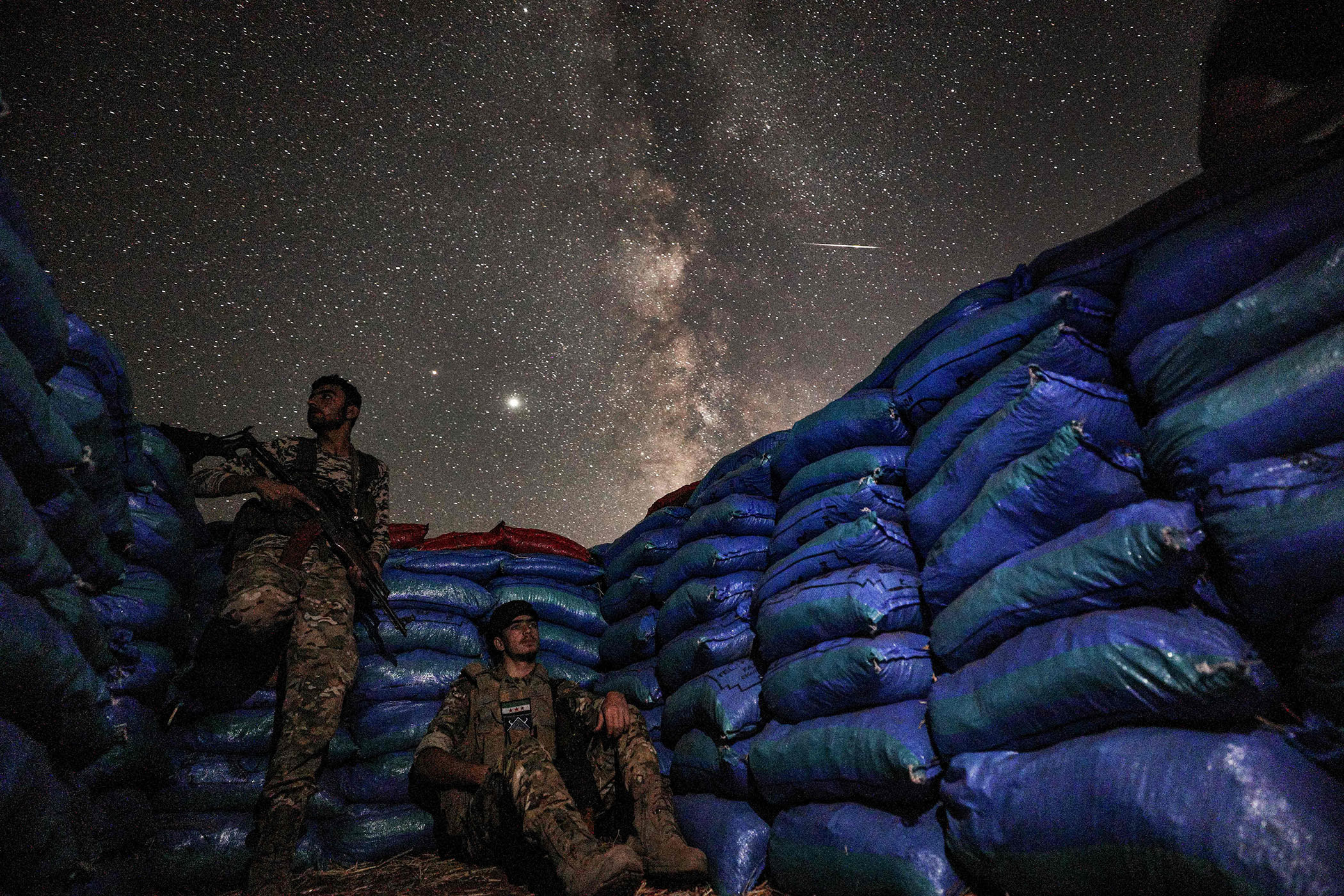 Two Syrian soldiers, one sitting on the ground and the other standing, are surrounded by sandbags that form a barrier; above them, the Milky Way galaxy and many stars are visible