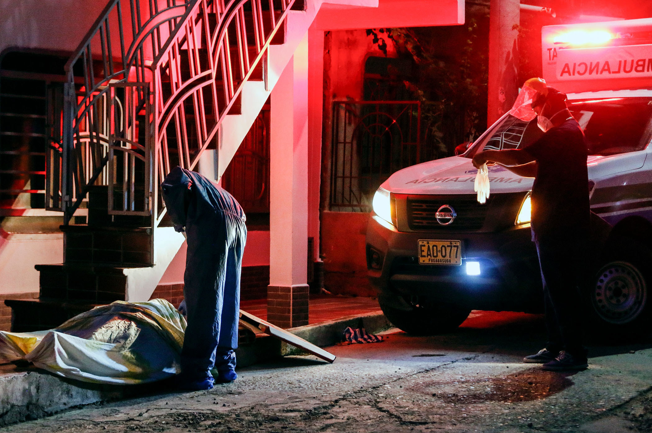 Two emergency medical workers stand near a sheet-covered body on the sidewalk; one worker puts on a glove while the other leans over the body