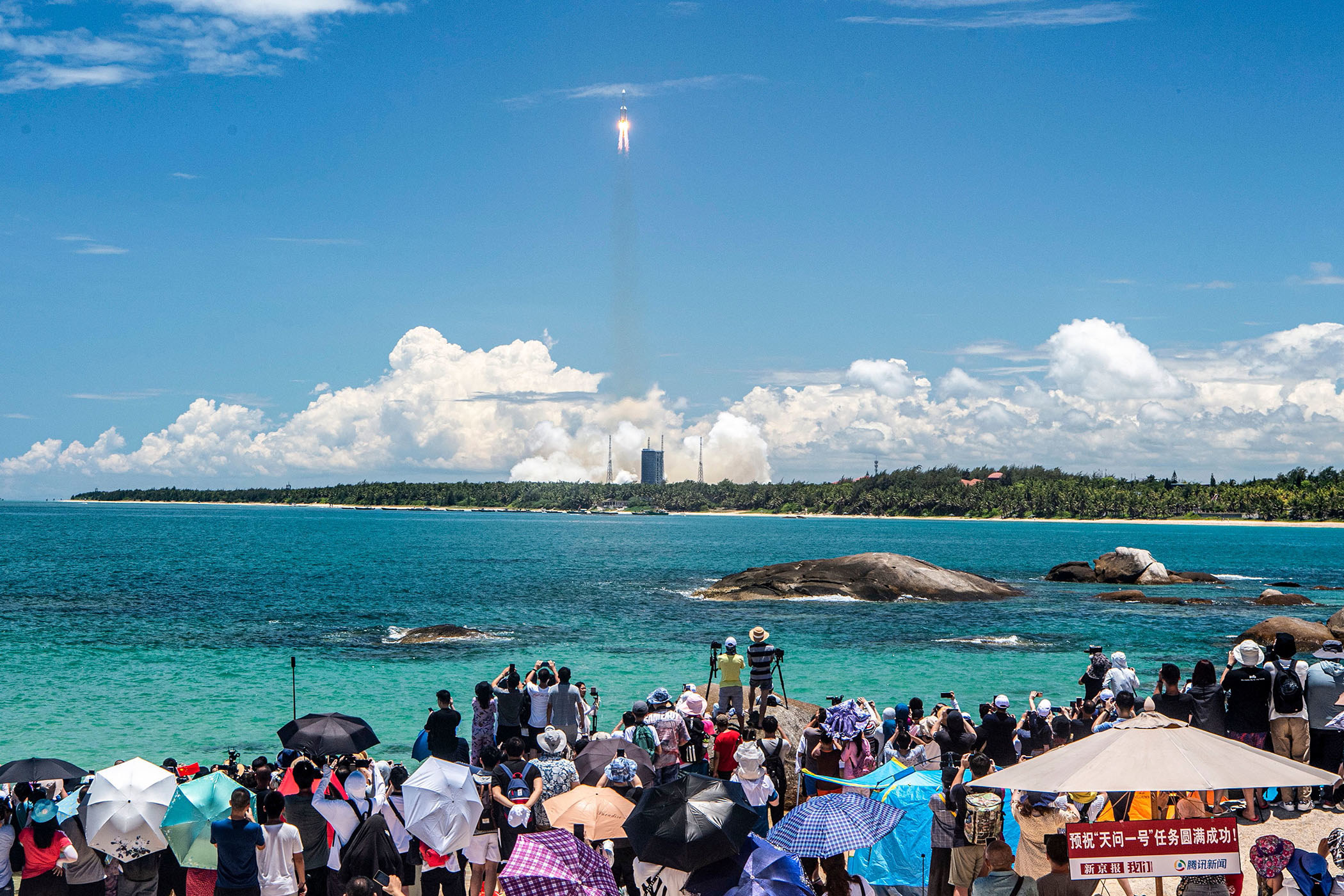 A crowd of people stand near the shore to watch a rocket launch in the distance; many of the people take pictures and shade themselves under umbrellas