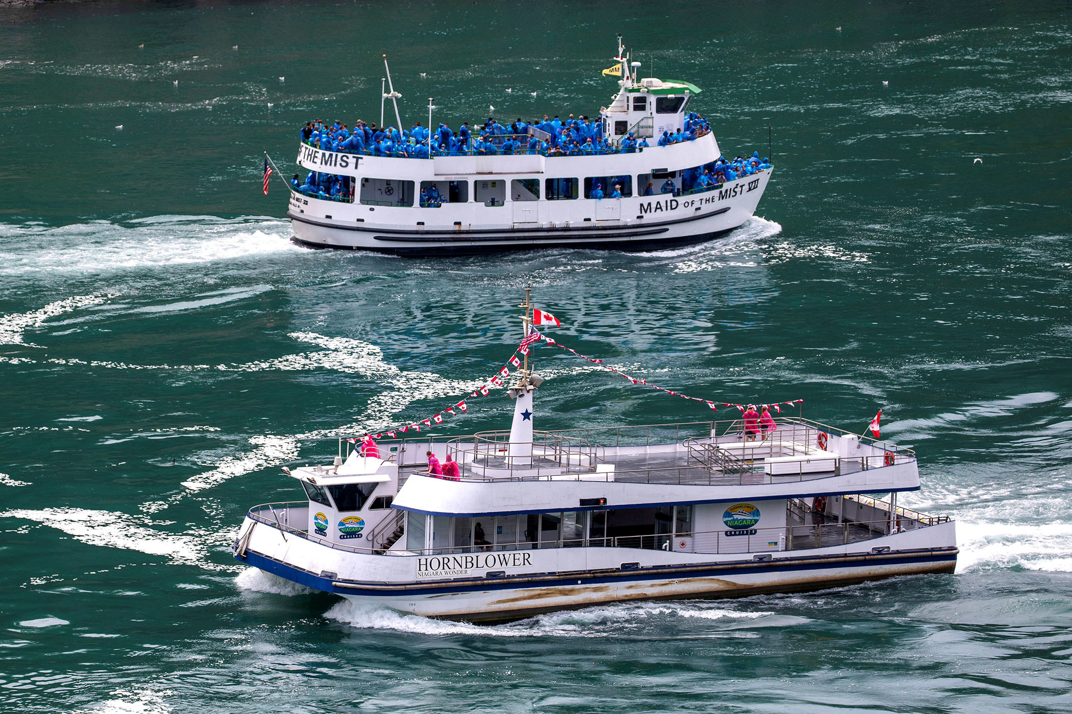 One boat in the foreground, named Hornblower, carries six passengers dressed in bright red raincoats; in the background, a tourist boat named &quot;Maid of the Mist&quot; carries several dozen passengers in blue raincoats