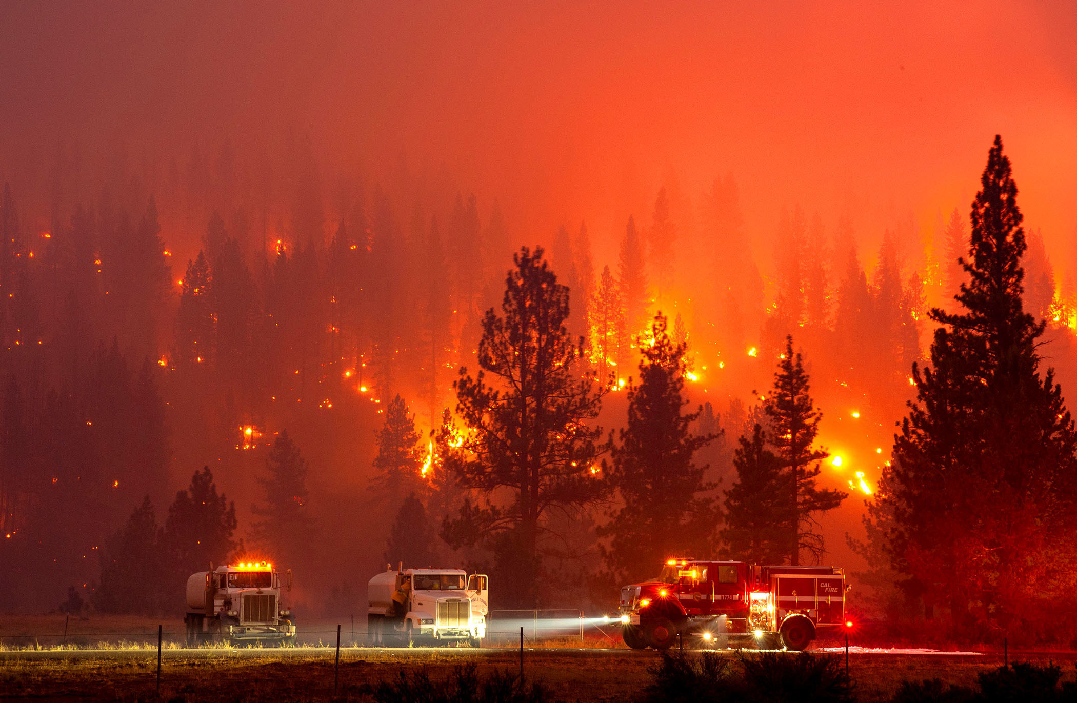 A fire burns through a hillside with trucks parked in a field below