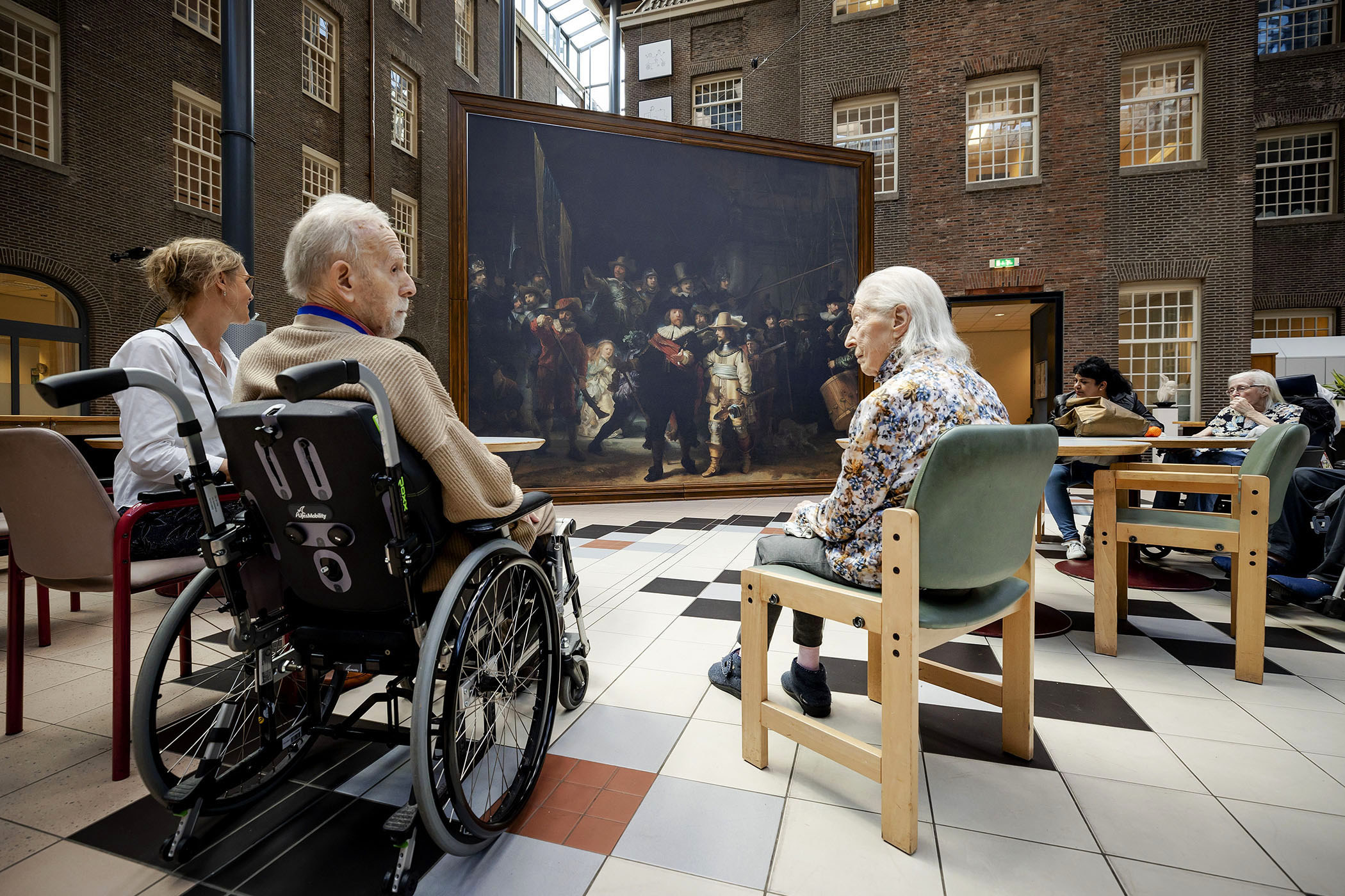 Older people sit in wheelchairs and in seats around a large Rembrandt painting,