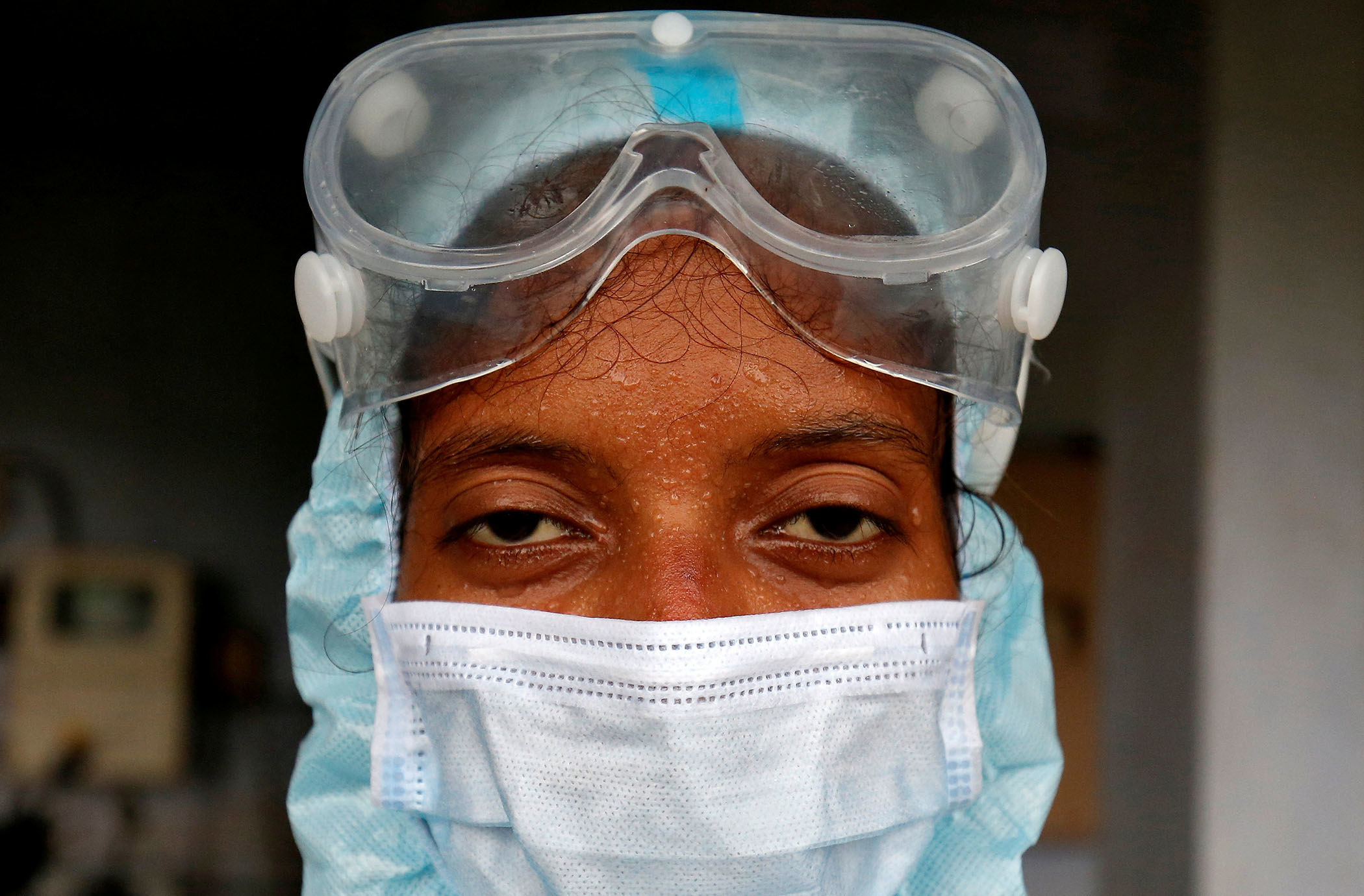 A healthcare worker, wearing a full-body protective gear, including a face mask and goggles, looks into the camera with tired eyes and a sweaty forehead