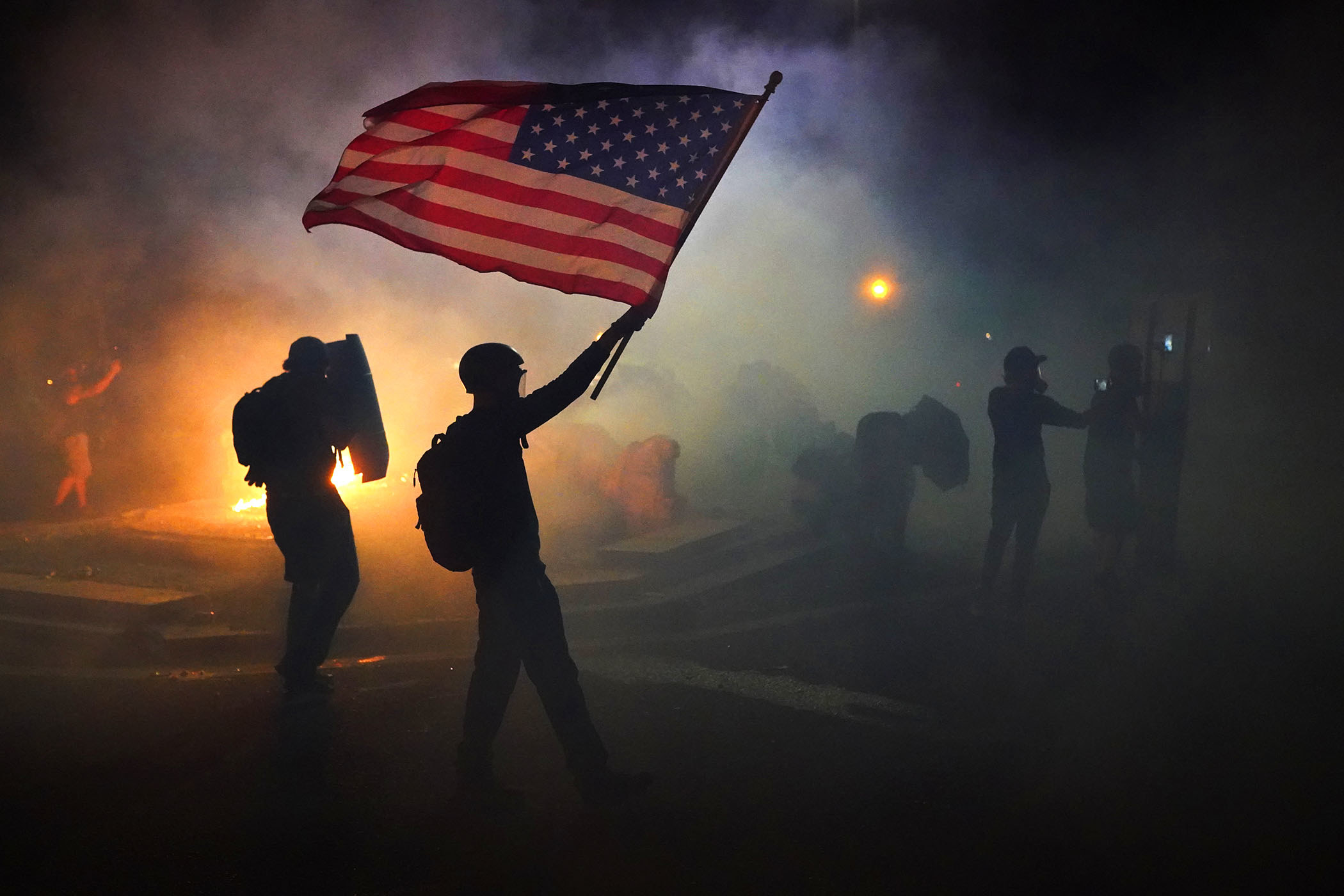 A demonstrator, backlit by a fire in the background, holds up a US flag amid clouds of tear gas