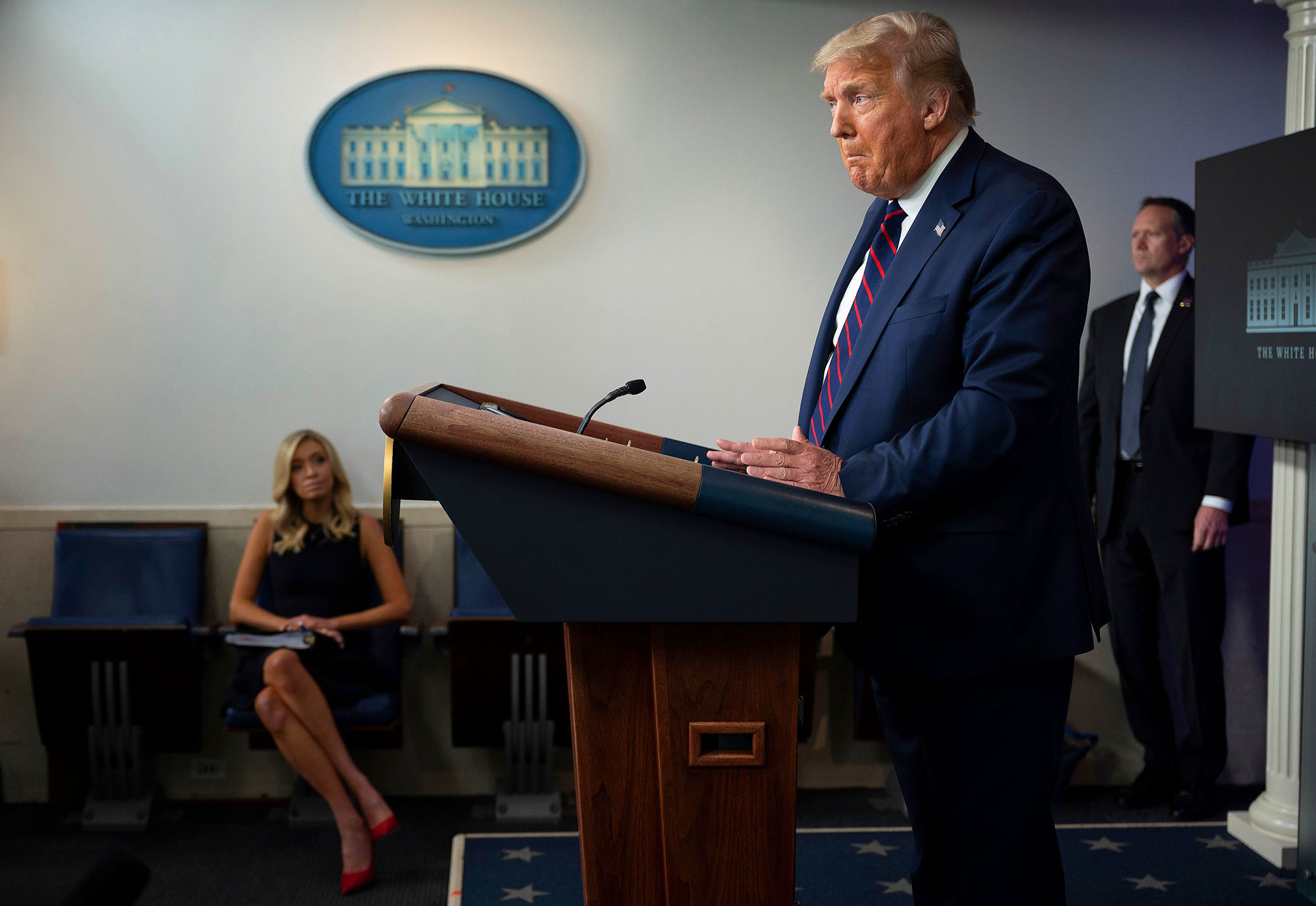 President Trump stands behind a lectern while press secretary Kayleigh McEnany sits in the background