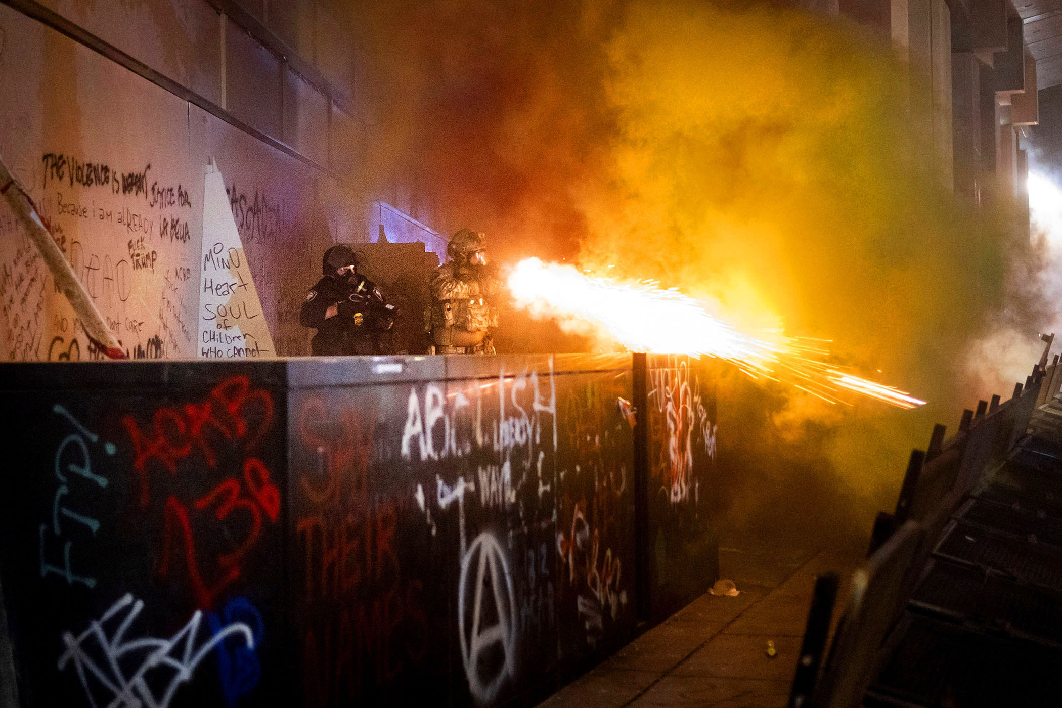 Standing behind a barrier covered in graffiti, a federal officer dressed in camouflage fires an explosive-looking munition
