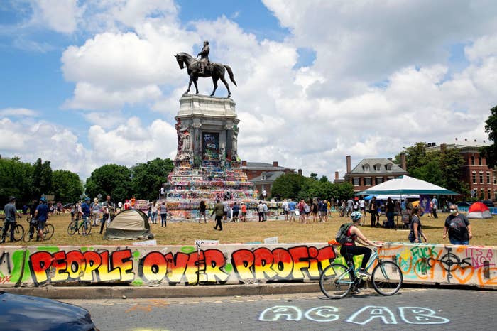 Graffiti reading "People Over Profit" is on a small barrier near grass surrounding the enormous statue of Robert E. Lee riding a horse. People surround the statue on bikes and in tent as the sun shines through clouds.
