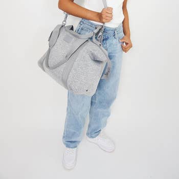 Model wearing the duffle bag on their shoulder using the long shoulder strap with a zipper across the top and shorter straps on the bag in heather grey