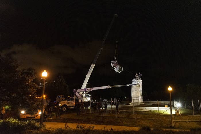 A crane removes a Christoper Columbus statue from its plinth amid nighttime darkness.