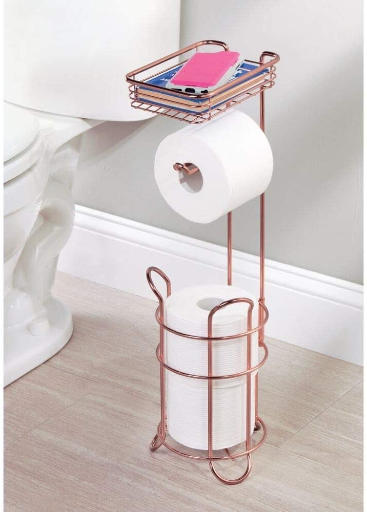 A toilet paper holder with one roll ready for use and two stacked for later use with a phone resting on top of the caddy
