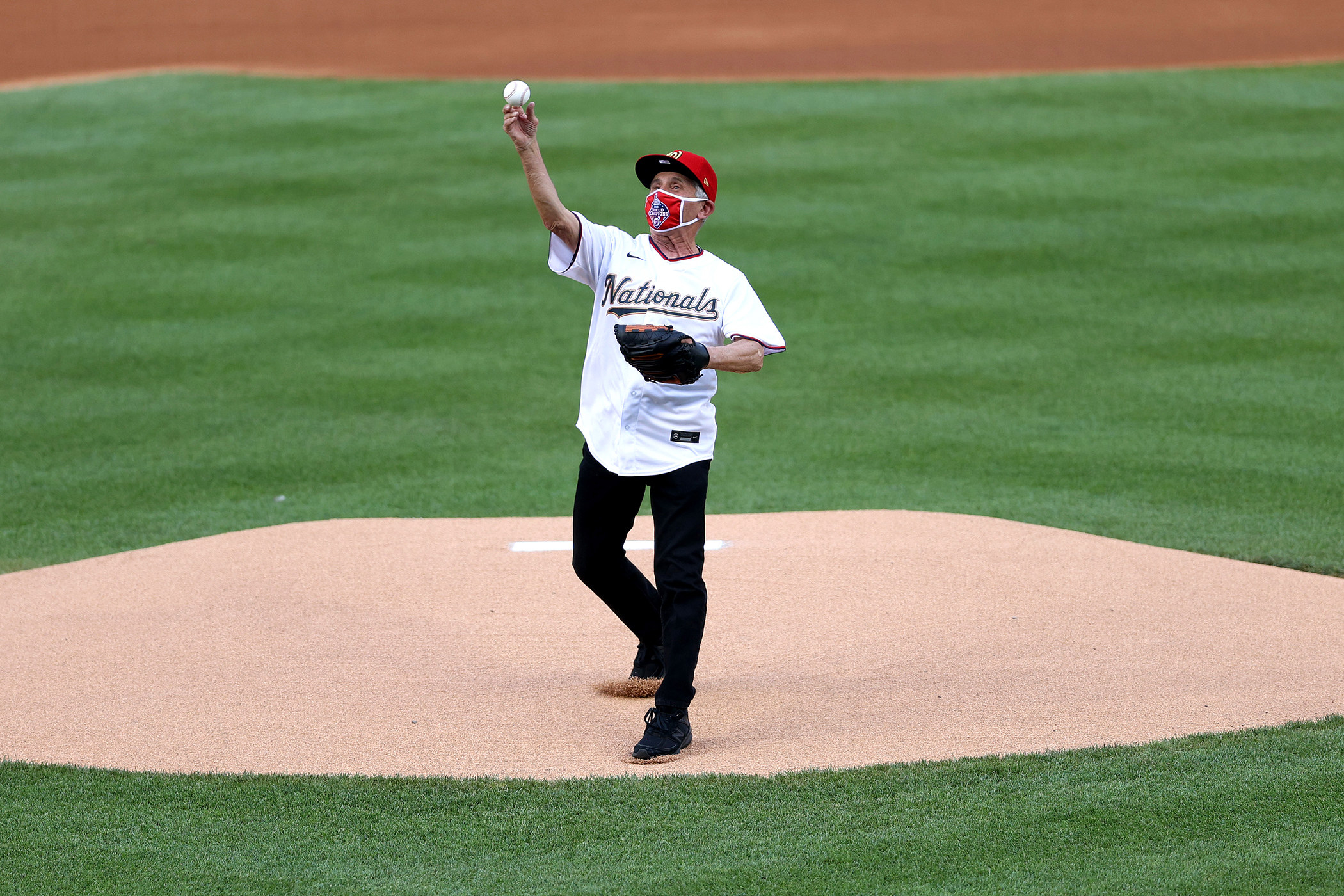 Anthony Fauci, wearing a Washington Nationals–branded cap, face mask, and jersey, stands on a mound and throws a baseball