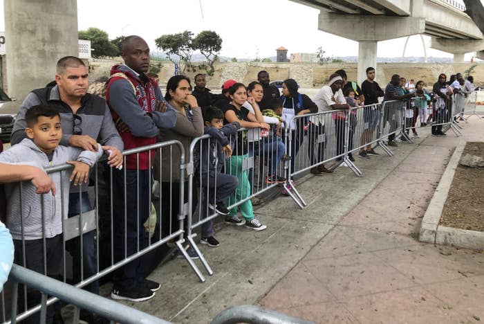 Dozens of people, many with sad expressions and arms crossed, wait behind a fence