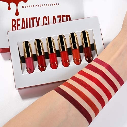 Six lipglosses are swatched on an arm in bars and the product is in its packaging
