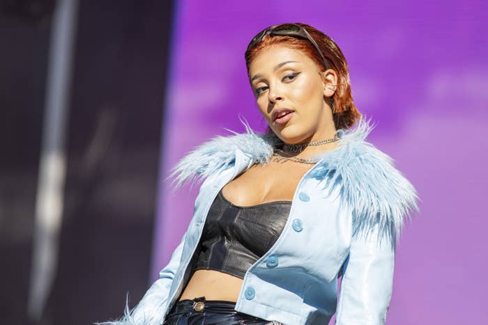 Doja Cat wears a leather bra, a jacket with a fluffy collar, and sunglasses on her head as she looks off camera while performing on stage.