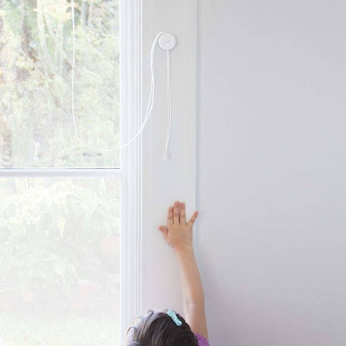 A child stretching up to reach the blind cord by the window without being able to reach it because the cord is wrapped around the round holder 