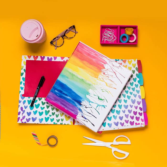 A binder with a rainbow water colour painting on the front There a set of a page dividers and a notebook underneath and a pen, scissors, tape, glasses, and coffee cup nearby