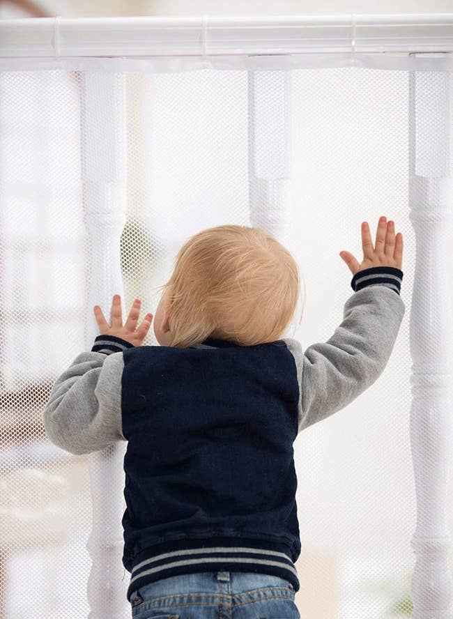 A baby standing up against a railing that is covered by the mesh gate, preventing them from slipping between the poles