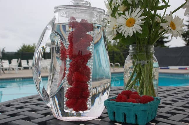 A clear fruit infuser pitcher with raspberries in the infuser 